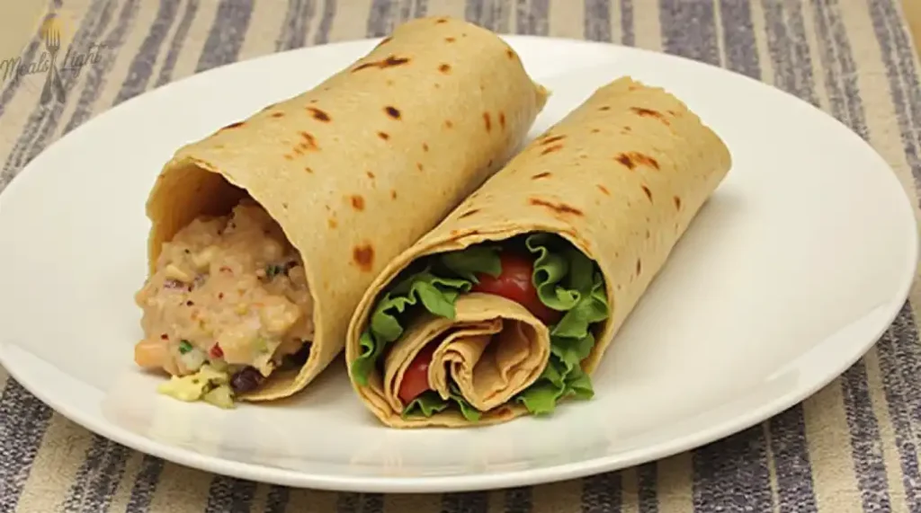 Two vegetarian wraps on a white plate. One wrap is filled with mashed beans and vegetables, while the other contains lettuce, tomatoes, and sliced fillings.